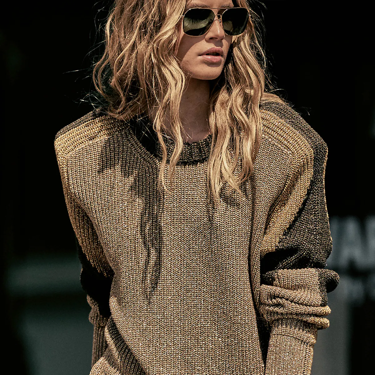Donna bionda con occhiali da aviatore, maglione a maglia grossa dorata e scintillante sulle spalle, maniche a blocchi neri, su sfondo scuro.