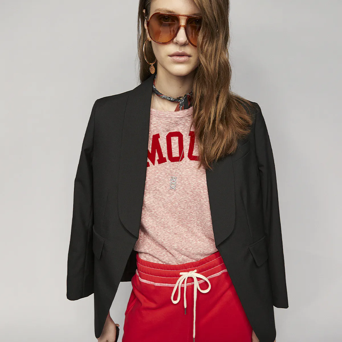 Donna con occhiali da aviatore, giacca da tailleur nera su una T-shirt rosa mélange stampata in rosso e pantaloni da jogging rossi