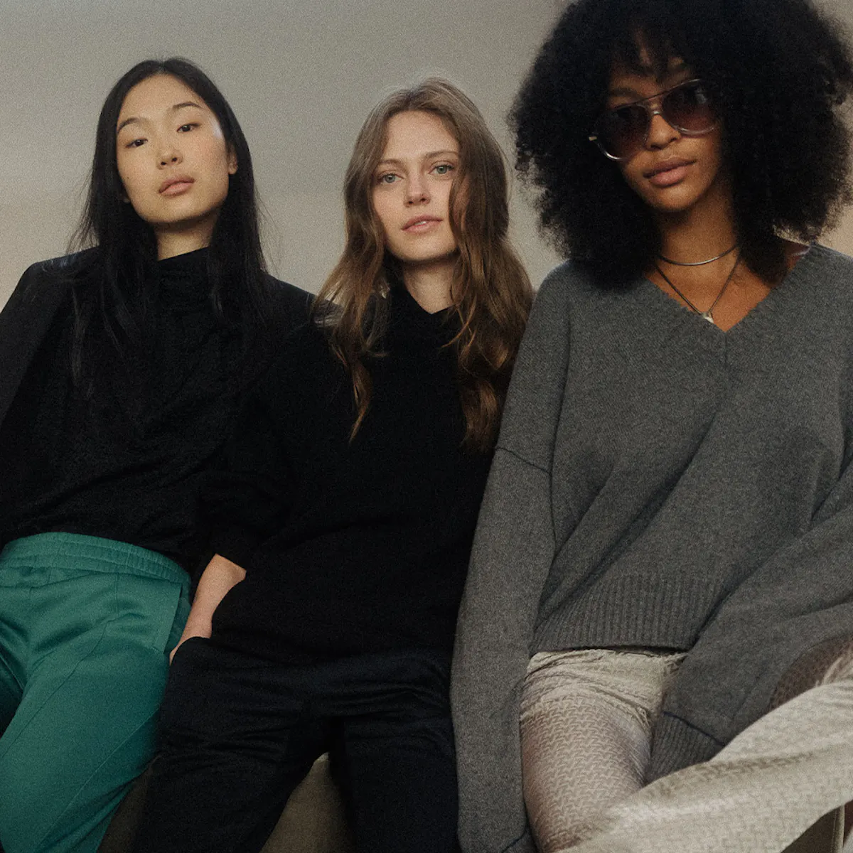Three women seated side by side facing the camera, two in black, the third in a grey V-neck jumper and tinted glasses.