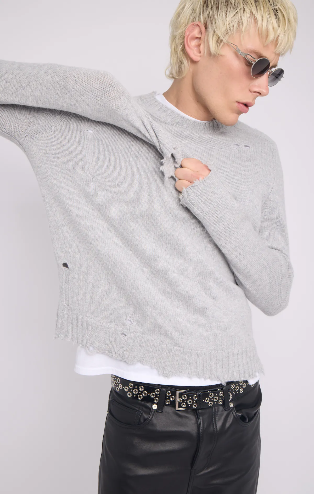 Un homme blond à lunettes rondes tire sur la manche de son pull gris clair, troué et effiloché aux poignets et à l’ourlet, porté sur un tee-shirt blanc avec une ceinture cloutée et un pantalon de cuir noir.
