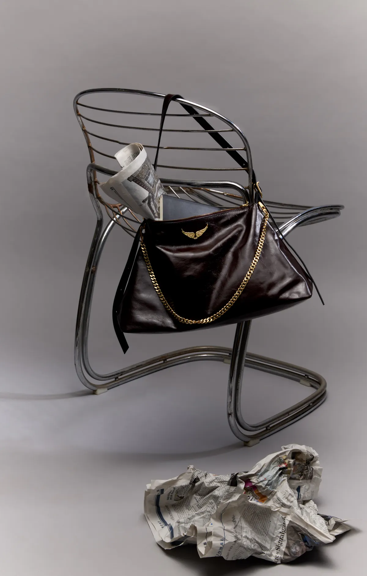 A Jim bag in soft dark brown leather, with gold chain and wings, hanging from the back of a chrome metal chair, a crumpled newspaper on the floor.