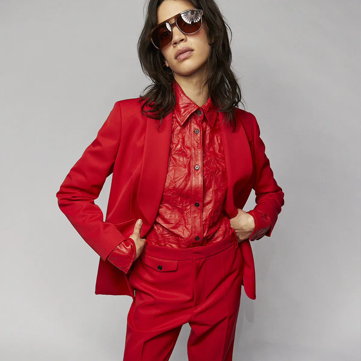 A woman in oversized sunglasses, hands on hips, wearing a red trouser suit over a red crinkled leather shirt.