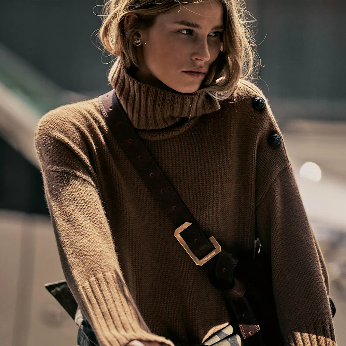 Donna con maglione a collo alto in maglia di cashmere color cammello, bottoni sulla spalla, una tracolla in pelle con fibbia dorata di traverso sul busto.