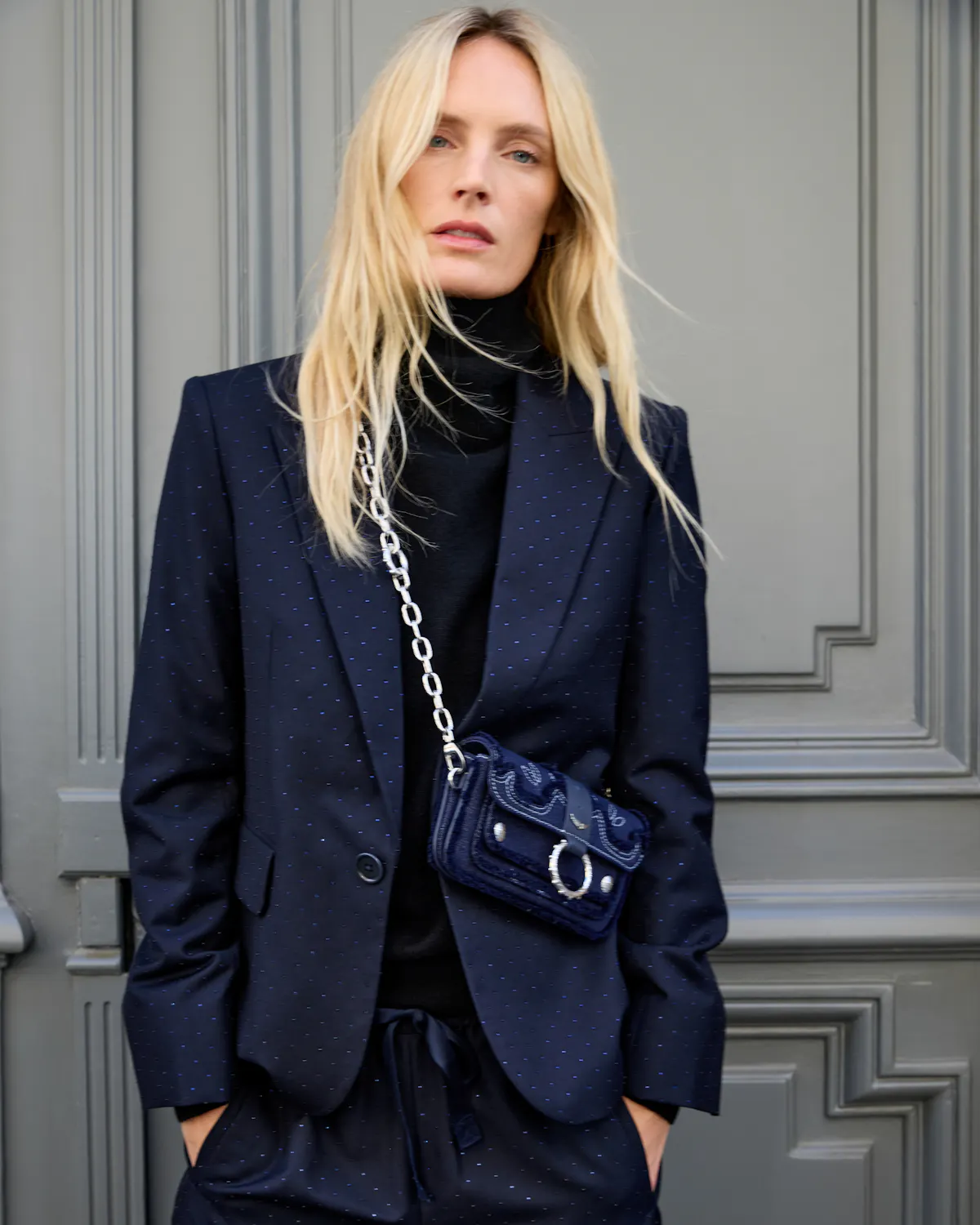In front of a grey panelled door, a blonde woman in a flecked navy suit and black roll-neck wears a Kate Wallet Denim bag crossbody, in navy denim with western topstitching and frayed edges, with a silver ring and chain.