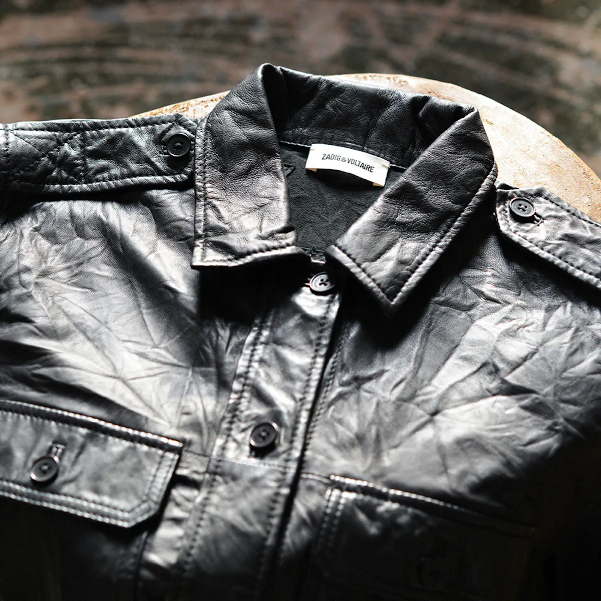 Camicia in pelle stropicciata nera appoggiata su uno sgabello, colletto, spalline e tasche con patta abbottonate, l'etichetta Zadig&Voltaire sul colletto, in luce radente.