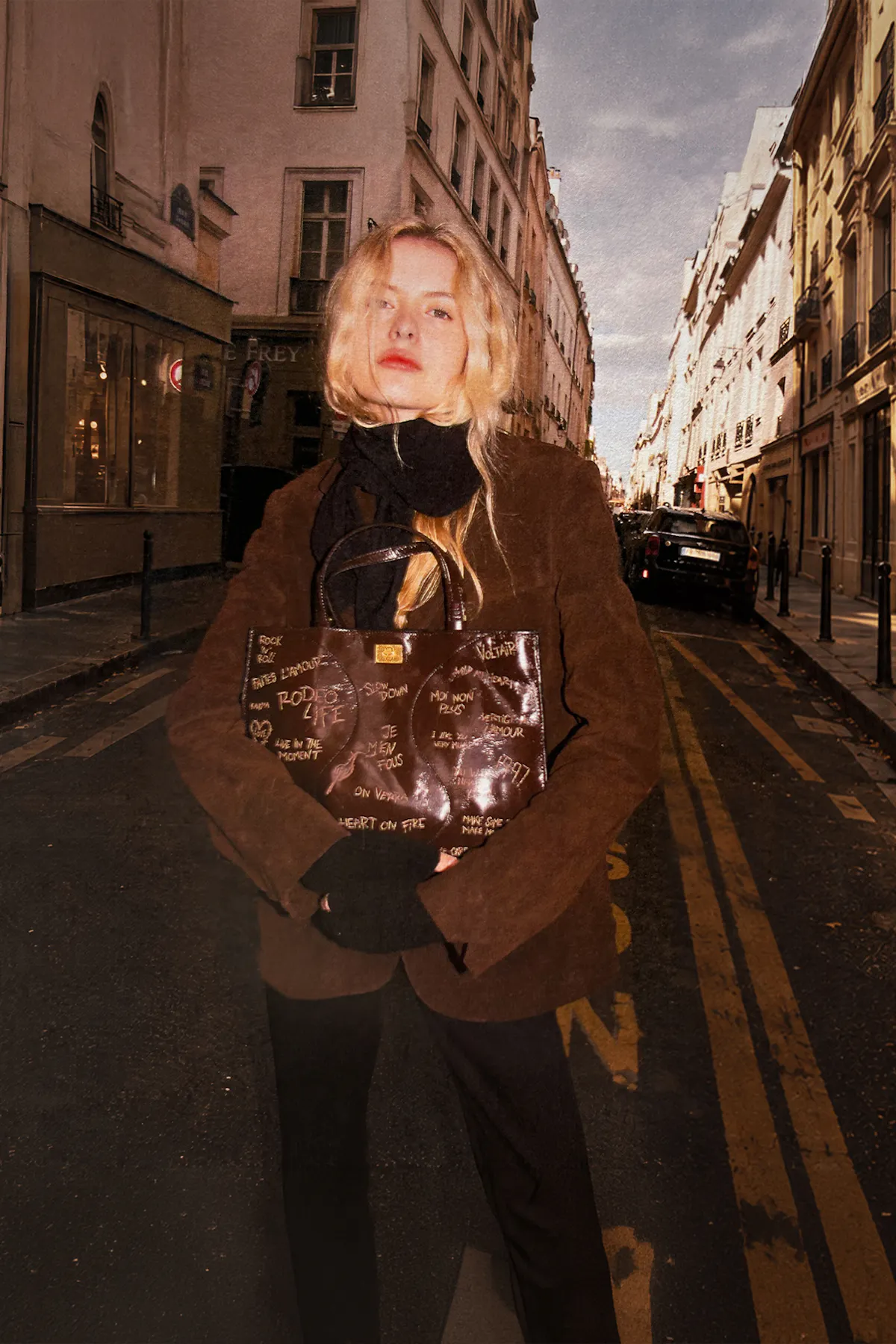 On a Paris street, a blonde woman in a brown suede jacket and black scarf holds close a brown patent leather bag covered in graffiti lettering.