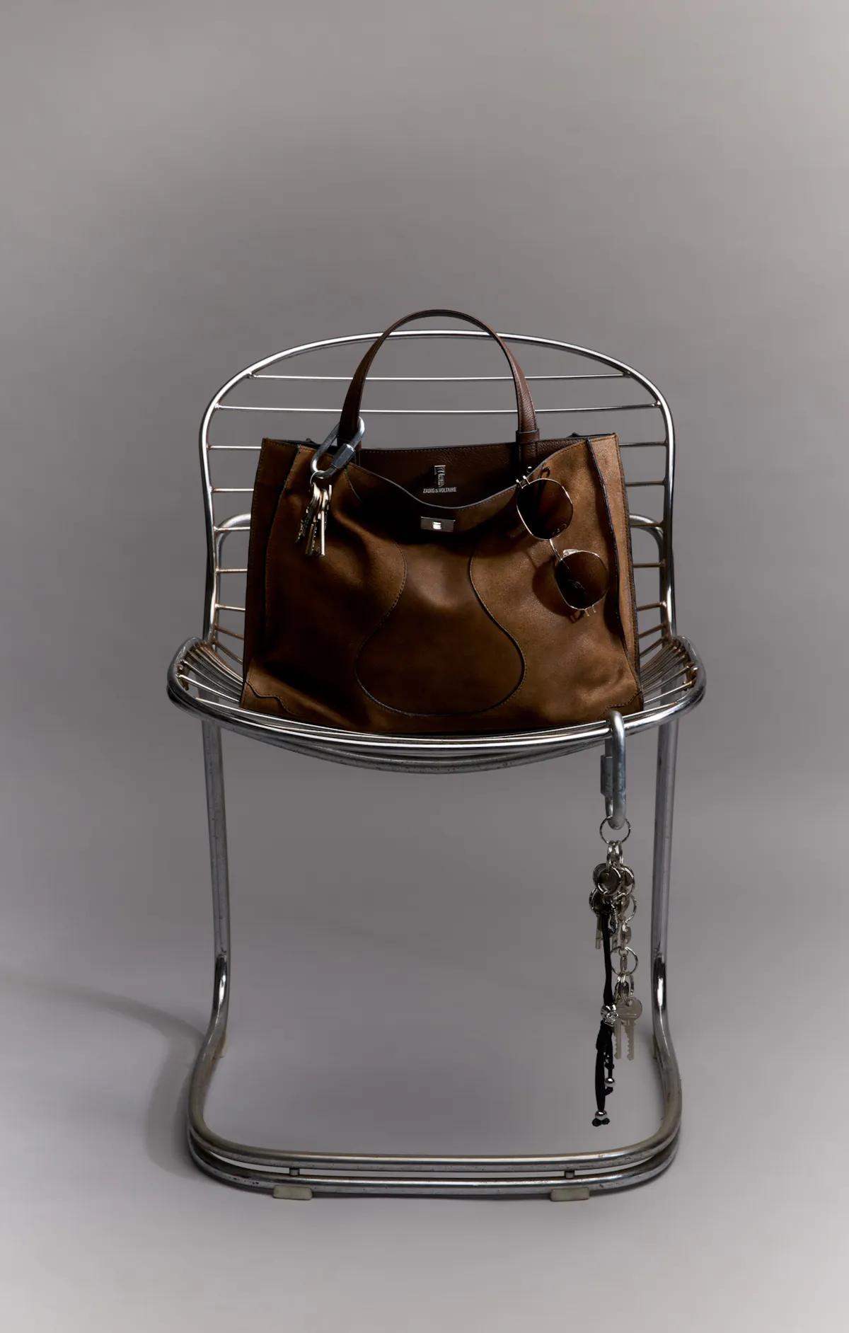 A Jack bag in brown suede with a clasp, set on a chair of chrome-plated metal tubing, with a bunch of keys and sunglasses hanging from its handles.