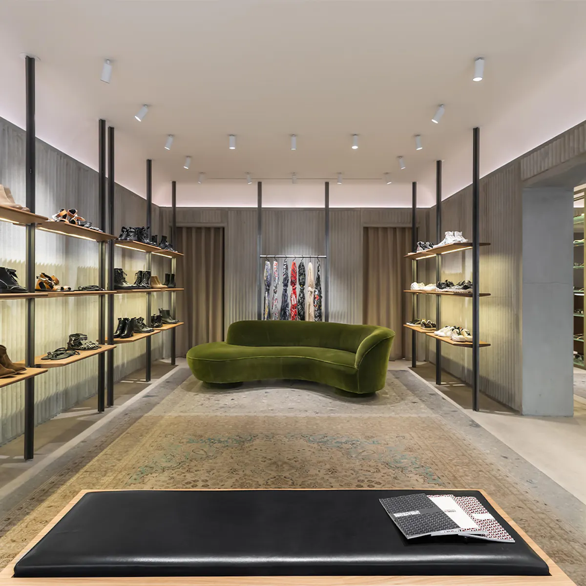 Cambon boutique: curved green velvet sofa on an antique rug, ribbed concrete walls, shoe shelves and a black leather bench.