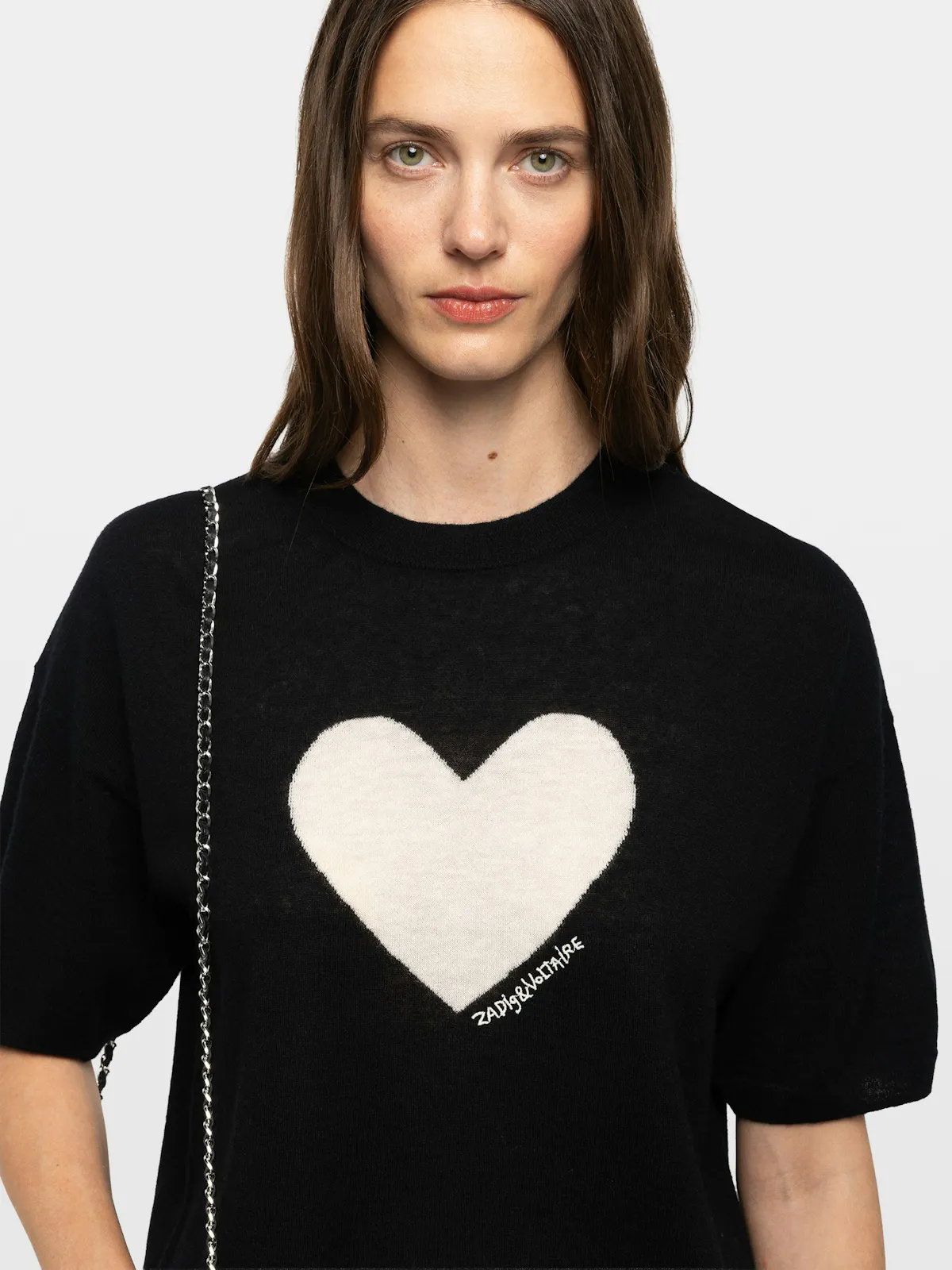 Une femme brune porte un pull noir à manches courtes, un grand cœur écru tricoté sur la poitrine.