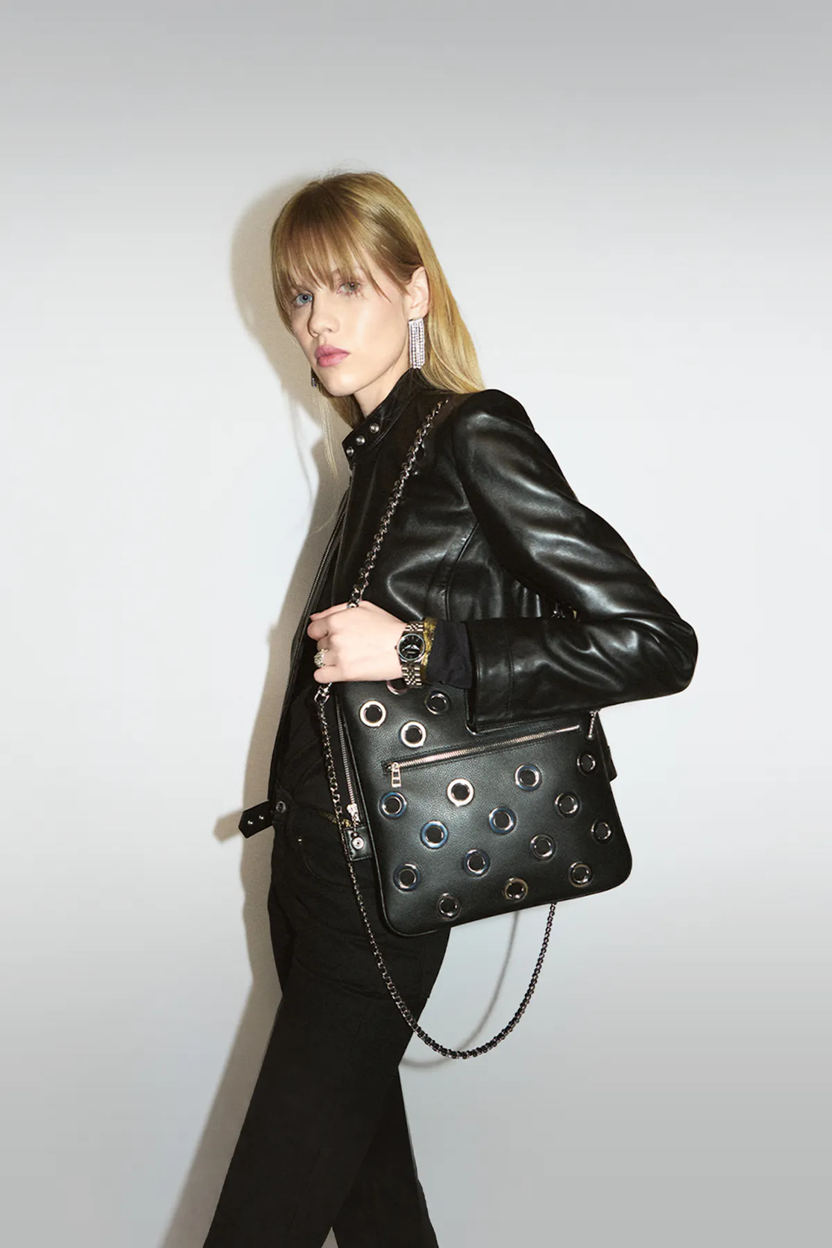 A woman in a black leather jacket wears a black Rock clutch studded with silver eyelets on her shoulder, on its chain.