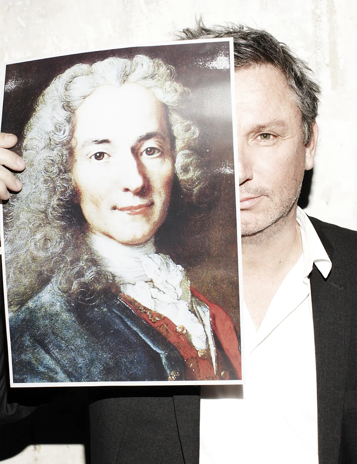 Thierry Gillier in giacca nera e camicia bianca, che tiene davanti a metà del viso la riproduzione di un ritratto dipinto di Voltaire