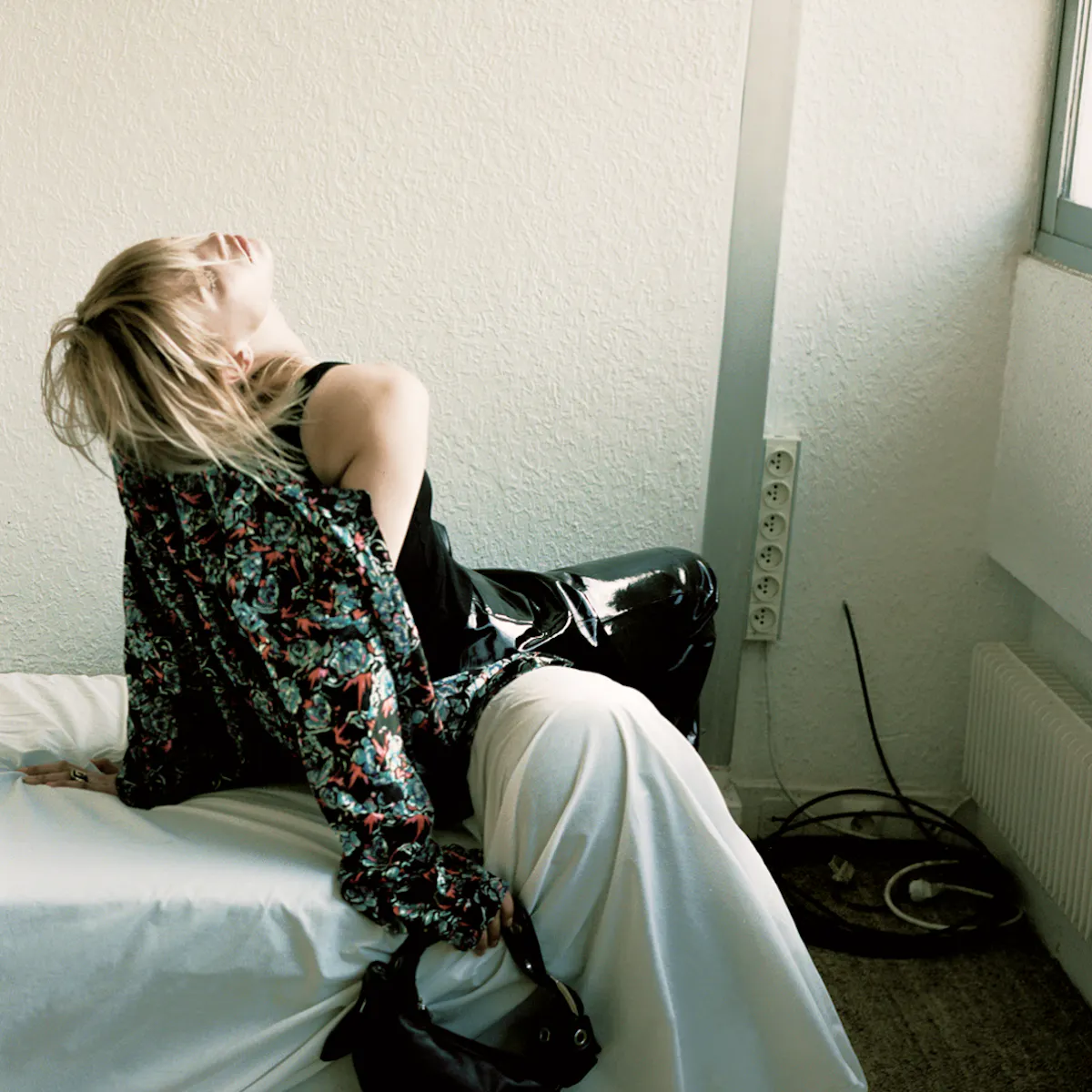 Donna bionda distesa all’indietro su un letto vicino a una finestra, giacca a fantasia floreale, pantaloni bianchi, borsa nera posata accanto a lei