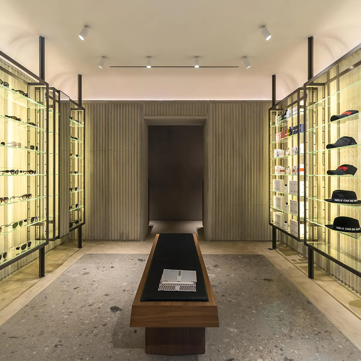 Room with lit glass display cases at the Cambon boutique, ribbed concrete wall and black leather bench.