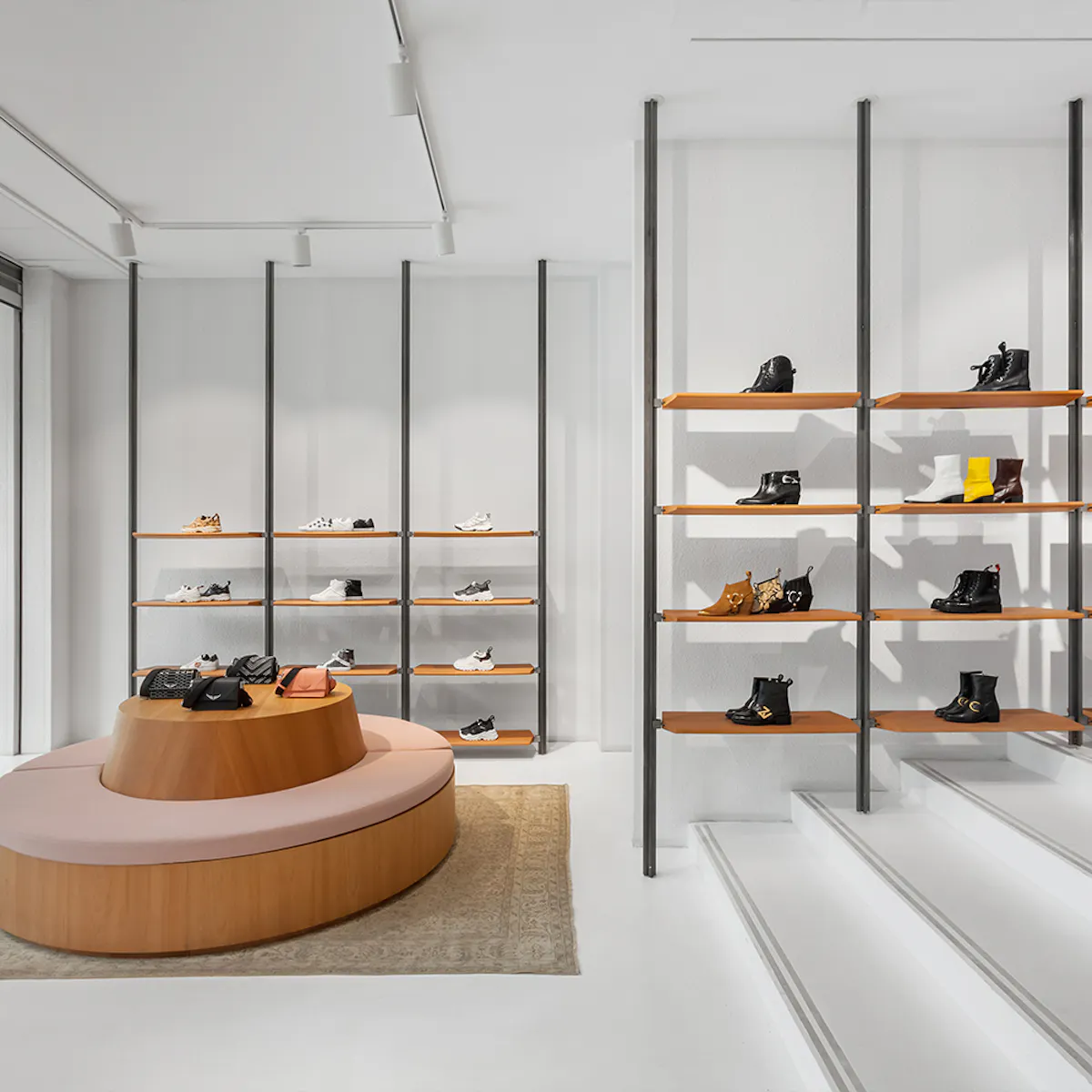 Wooden shelves on black uprights lined with ankle boots and trainers, a round wooden island with a pink banquette, and white steps.