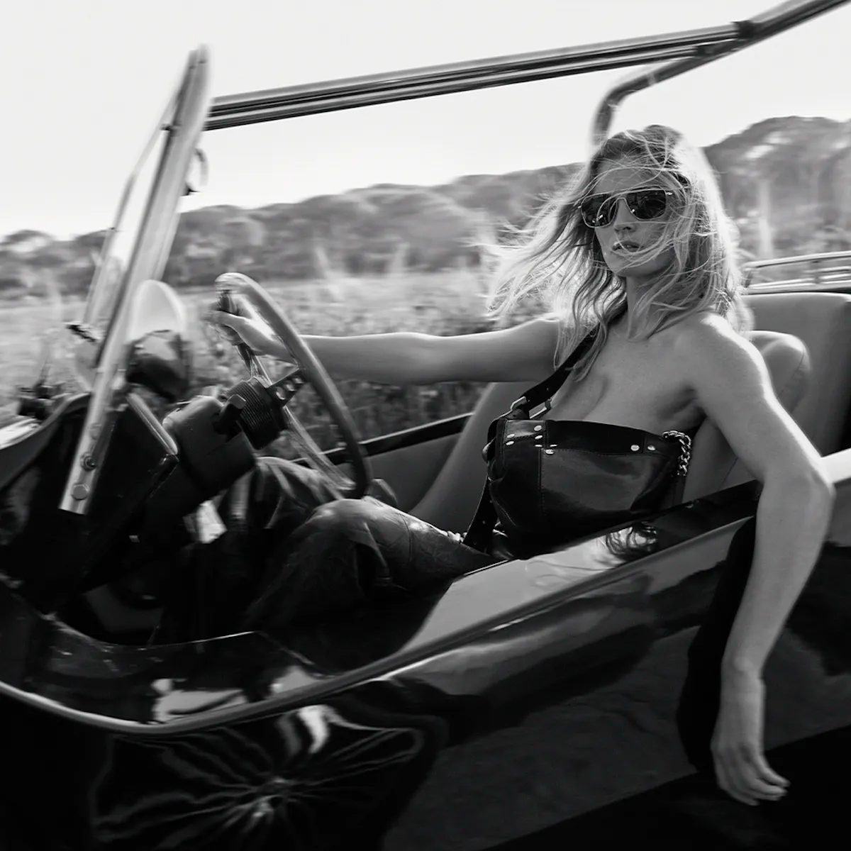 At the wheel of an open-top buggy, a woman in sunglasses, hair in the wind, a studded black leather bag worn crossbody.