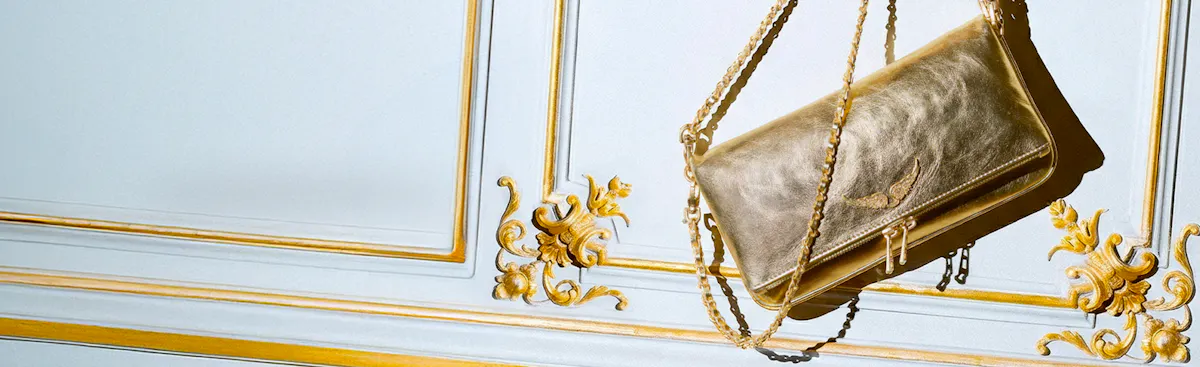 Rock clutch in metallic gold leather hanging by its chain in front of pale blue panelling with gilded mouldings.